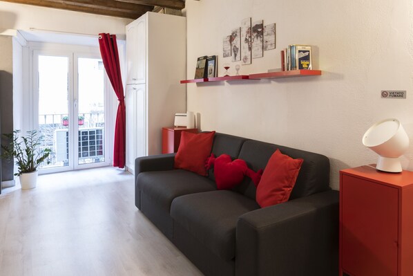A modern living room with a dark grey sofa, red cushions, a red cabinet with a lamp, and a bookshelf with books and framed pictures. - notaMi -  Casa Vittoria - Isola District (Milan)