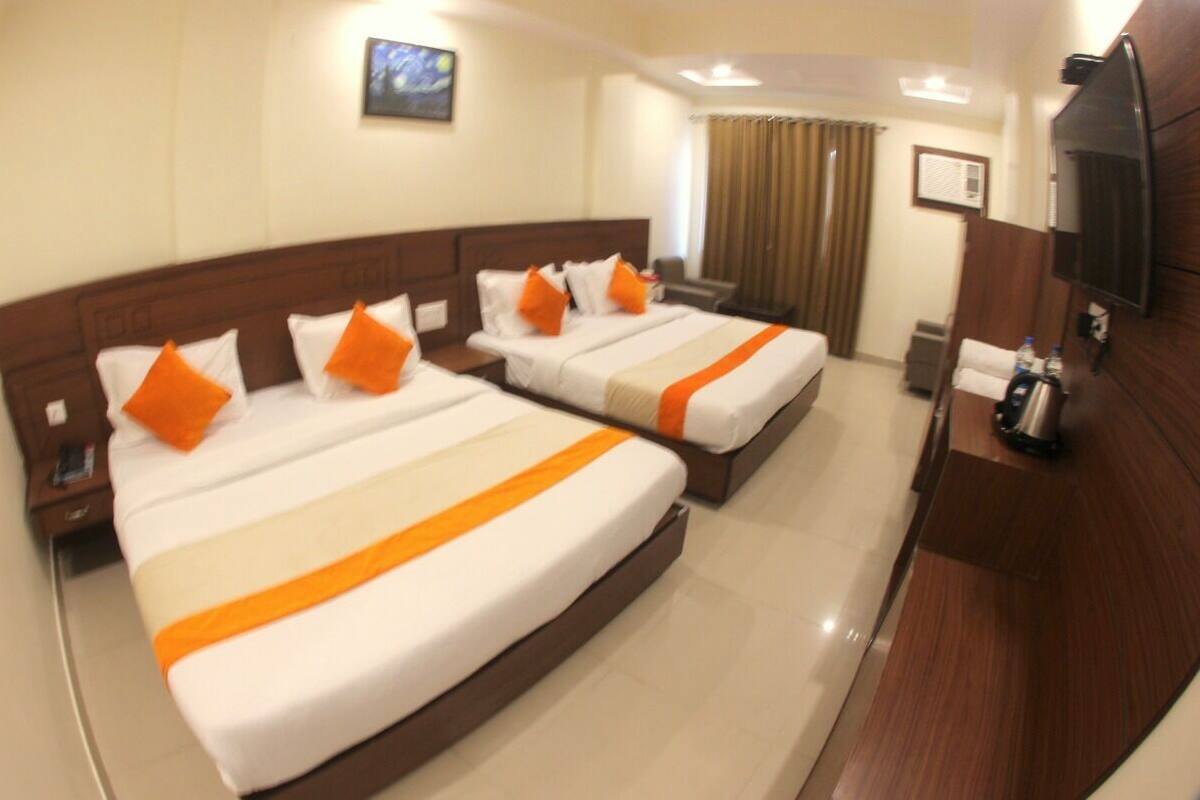 A hotel room with two beds, each with white bedding and orange accents, a wooden headboard, a flat-screen TV, and a small desk with a chair.