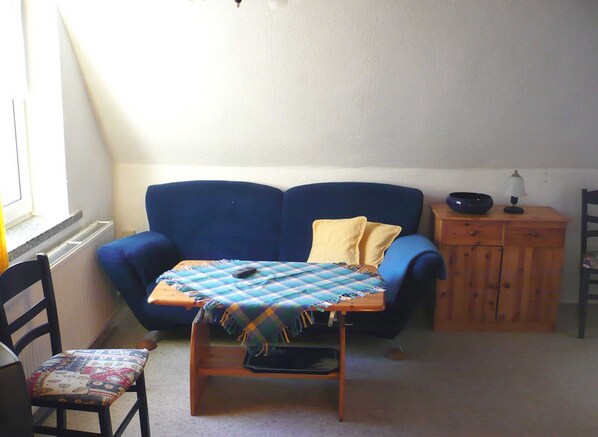 A room with a blue sofa, a wooden cabinet, a small table with a checkered tablecloth, and a chair with a patterned cushion. - Vacation apartment 2 (stairs) - Egons Hüsing (Insel Hiddensee)