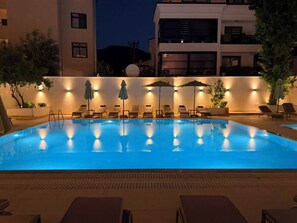 An outdoor swimming pool with lounge chairs and umbrellas, surrounded by a low wall and buildings in the background.