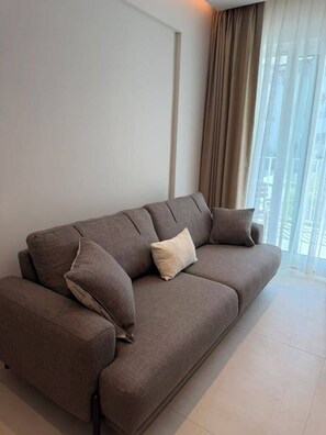 A modern living room with a sofa, a window with sheer curtains, and a view of the outside. - Pasahan Apart (Marmaris)