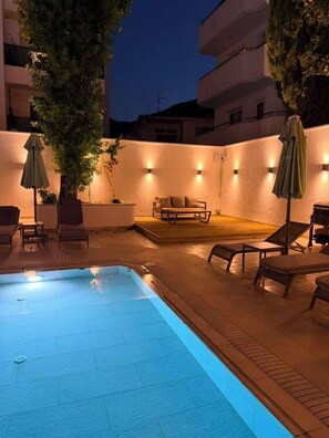 A swimming pool area with lounge chairs and umbrellas, surrounded by a low wall and a building. - Pasahan Apart (Marmaris)