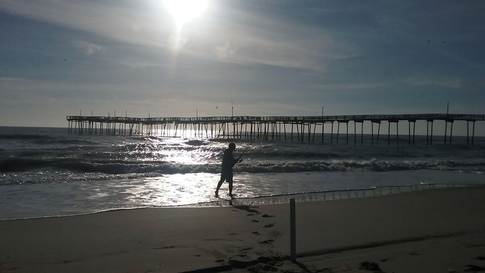 A beach scene with a pier extending into the ocean, a person fishing, and footprints leading to the water.