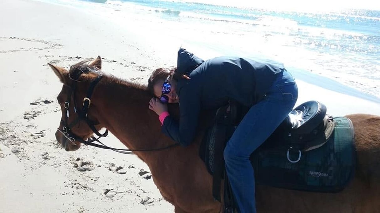 Une personne à cheval sur une plage, avec du sable et de l’eau en arrière-plan.