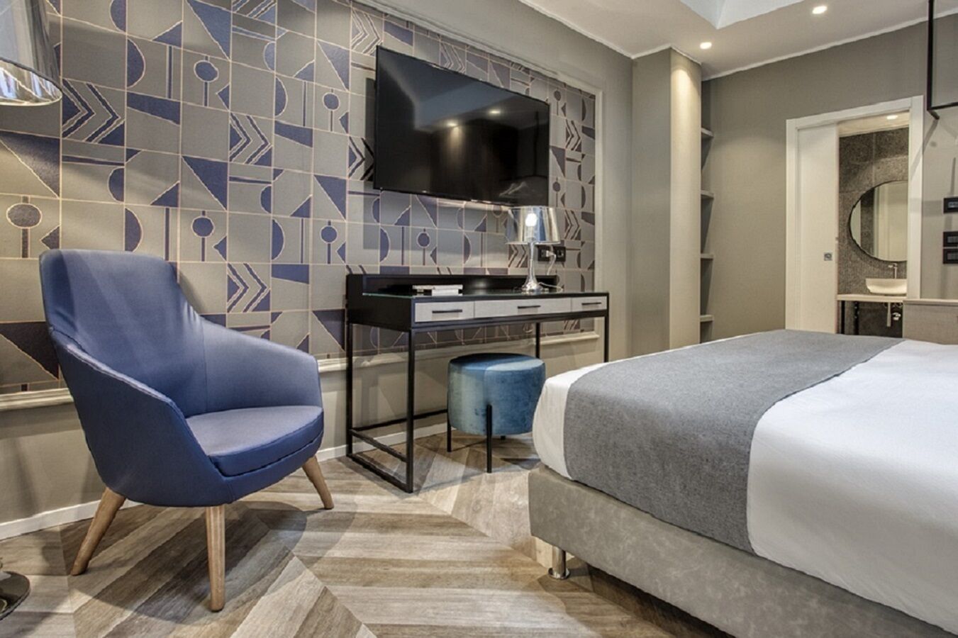A modern hotel room with a large bed, a desk with a flat-screen TV, a blue armchair, and a patterned wall.