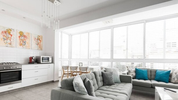 A modern kitchen with white cabinets, a grey sofa, and a dining area with a white table and chairs. - Beachside Apartment: Modern Comfort & Style (Tel Aviv-Yafo)