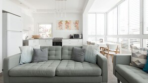 A modern kitchen with white cabinets, a dining area with a table and chairs, and a grey sofa with cushions. - Beachside Apartment: Modern Comfort & Style (Tel Aviv-Yafo)