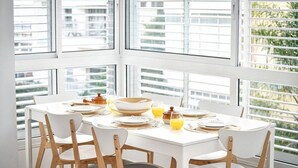A dining area with a white table set for four, surrounded by large windows with blinds, offering a view of buildings and greenery. - Beachside Apartment: Modern Comfort & Style (Tel Aviv-Yafo)