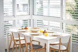 Un'area pranzo con tavolo bianco imbandito per quattro persone, circondata da ampie finestre dotate di persiane, che offre vista su edifici e vegetazione.
