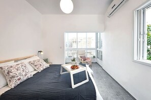 Un dormitorio moderno con cama, mesita de noche con bandeja, una silla y un ventanal con vistas a la ciudad.