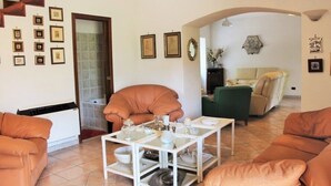 Ein Wohnzimmer mit einer Couch, einem Couchtisch und einem Sessel. An der Wand hängen gerahmte Bilder und im Hintergrund ist eine Treppe zu sehen.