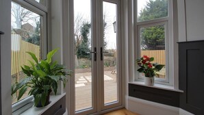 A room with a sliding glass door leading to an outdoor patio, a potted plant on the windowsill, and a small shelf with a flower pot. - Belmont Road (Broadstairs)