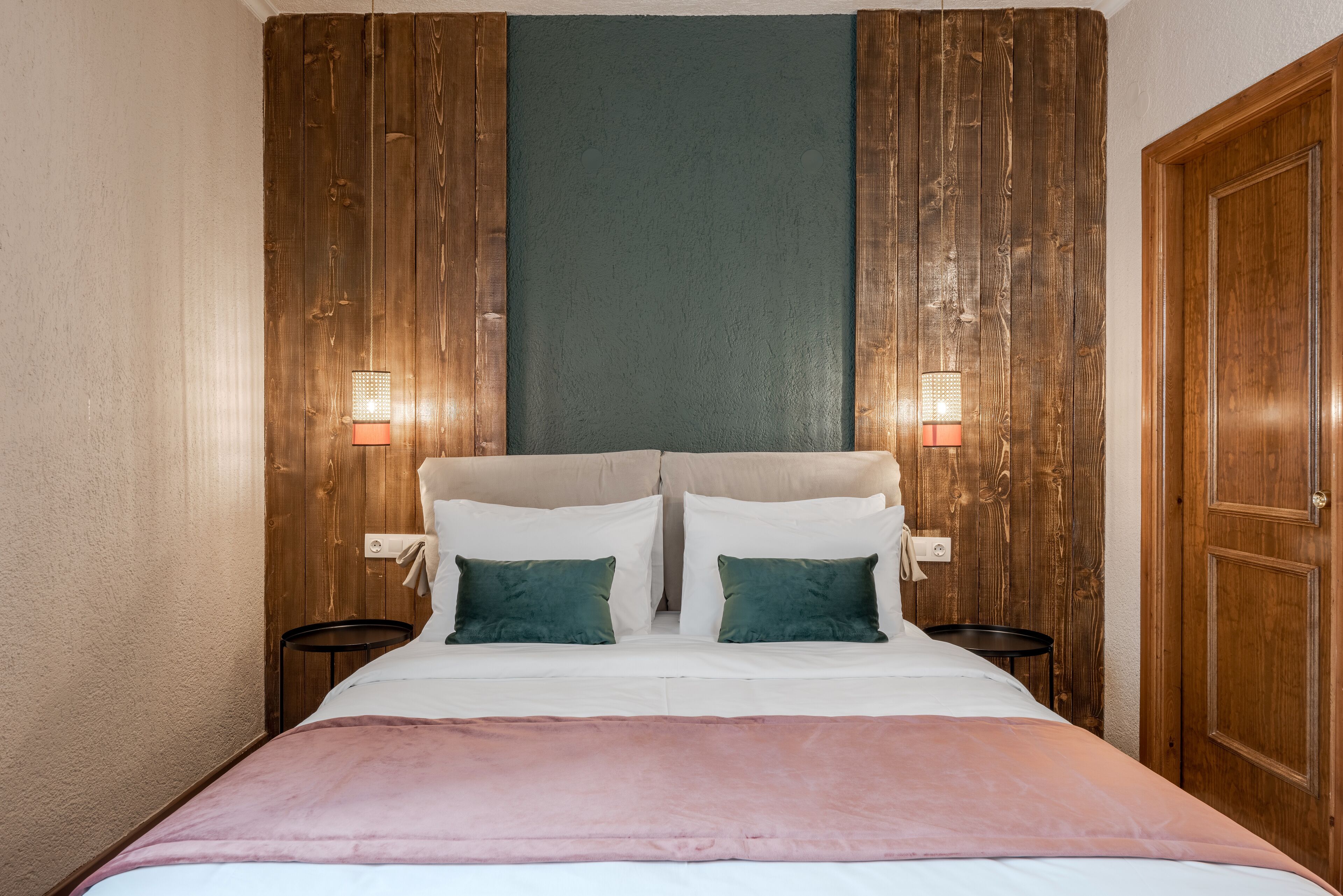 A double bed with a pink blanket, two green pillows, and two bedside tables with lamps.