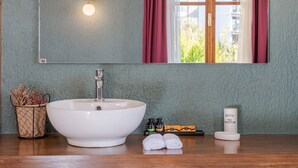 Um banheiro com uma pia grande e branca, um espelho, uma janela com cortinas vermelhas, uma pequena planta em uma cesta de arame e um frasco de sais de banho.