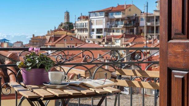 Uma mesa de madeira com um livro, uma xícara e um vaso roxo com uma flor, numa varanda com vista para uma paisagem urbana com edifícios de telhados vermelhos.