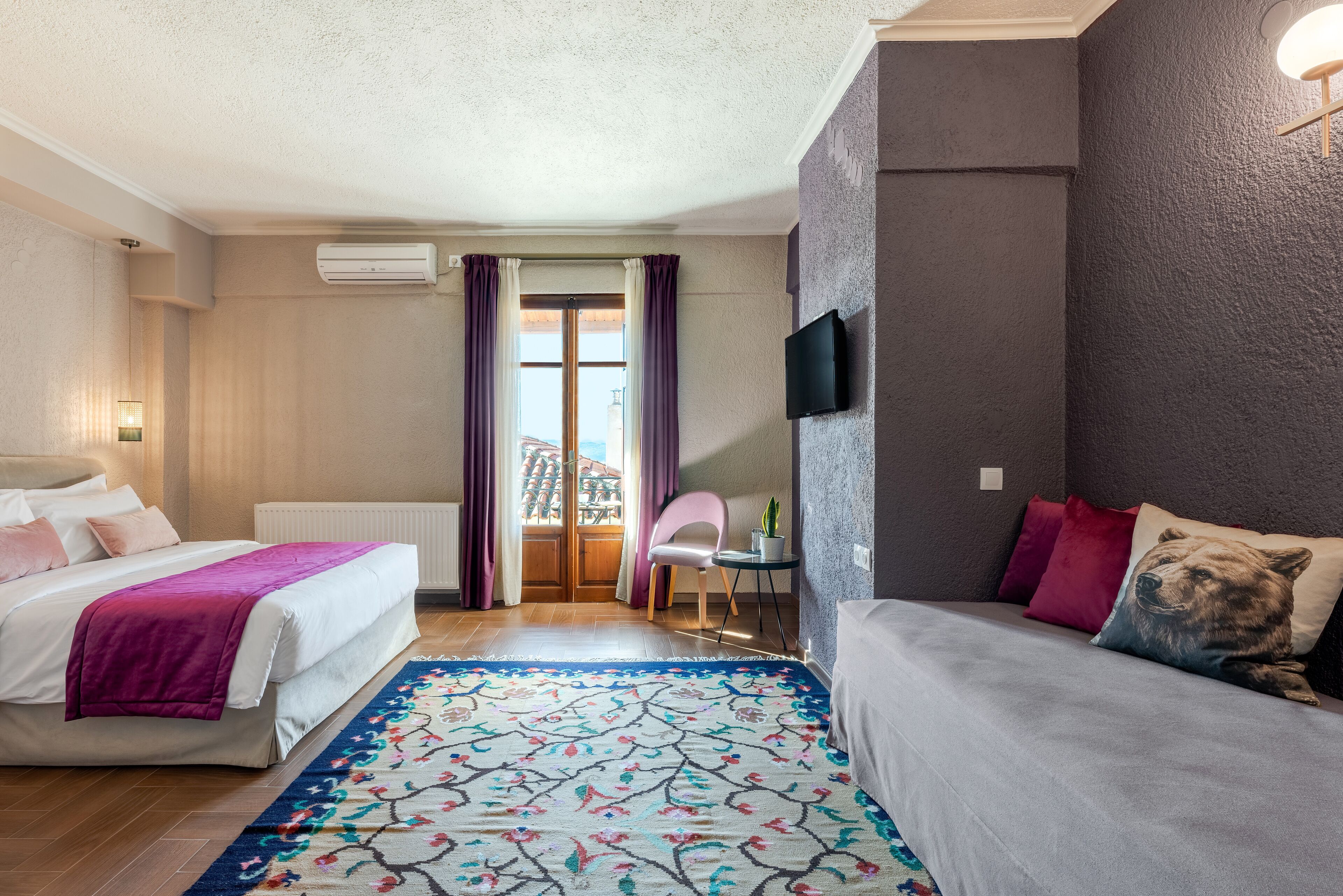 A hotel room with two beds, a TV, a window with curtains, a small table with a plant, and a colorful rug.