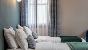 Quarto com duas camas, roupa de cama branca, almofadas estampadas, cortinas leves e um cobertor verde.