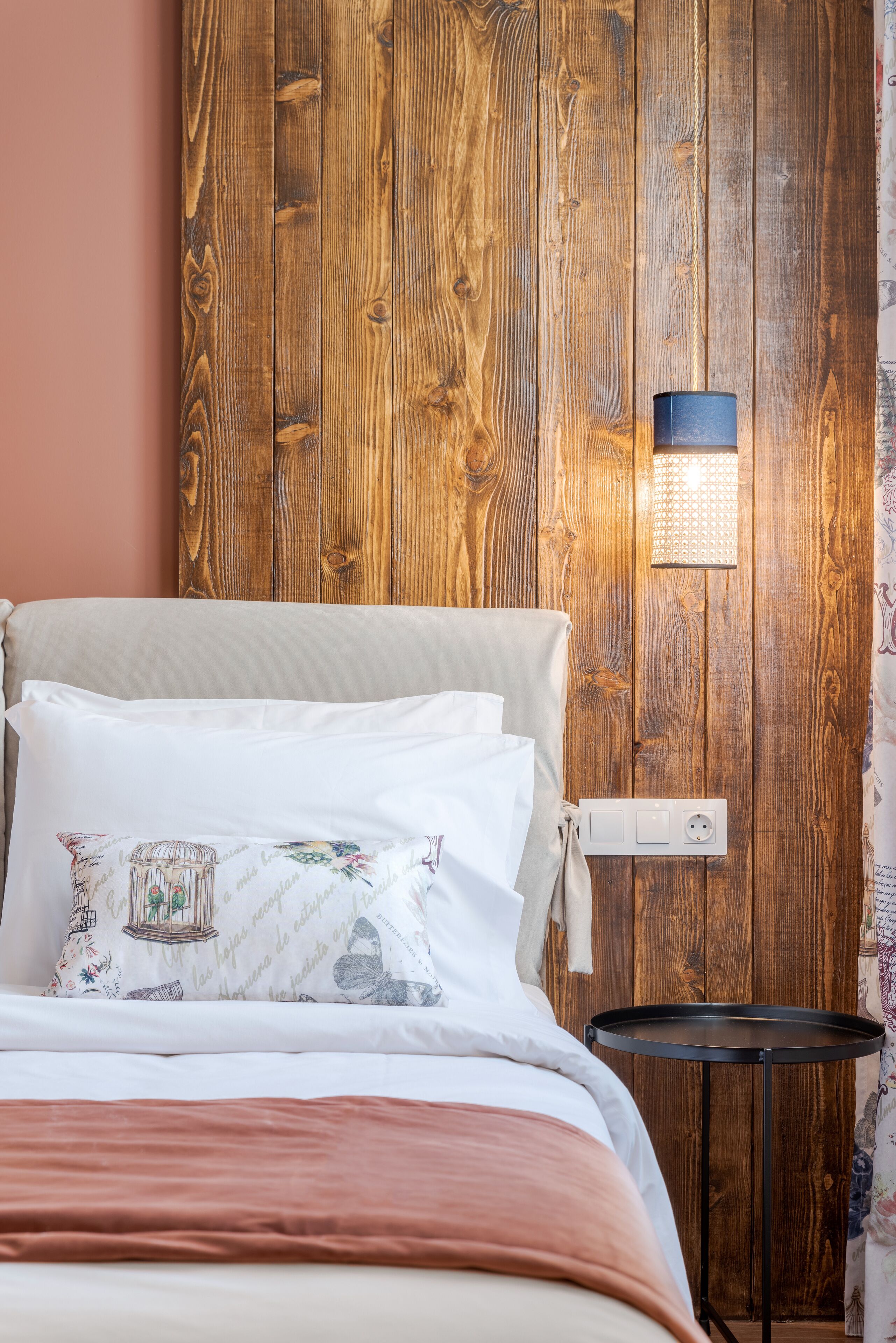 Een net opgemaakt bed met een bloemenkussen, een houten wandpaneel en een klein zwart tafeltje ernaast.