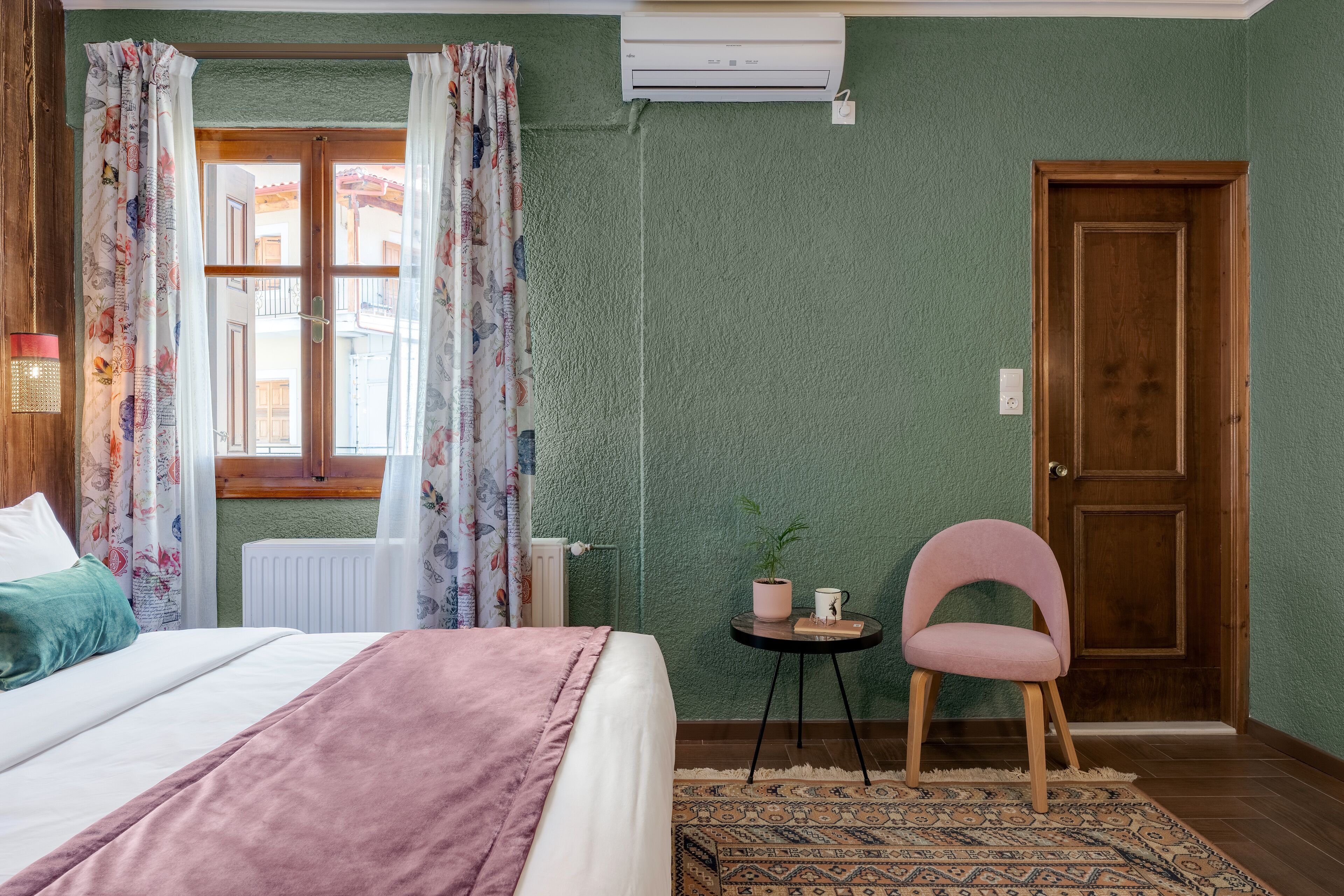 A bedroom with a bed, a chair, a small table with a plant and a cup, a radiator, and a door.