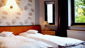 A hotel room with two single beds, a nightstand with a lamp, a wall-mounted light fixture, a window with a view of greenery, and a radiator. - Haus Martens Das Lister Stadthotel (Hannover)