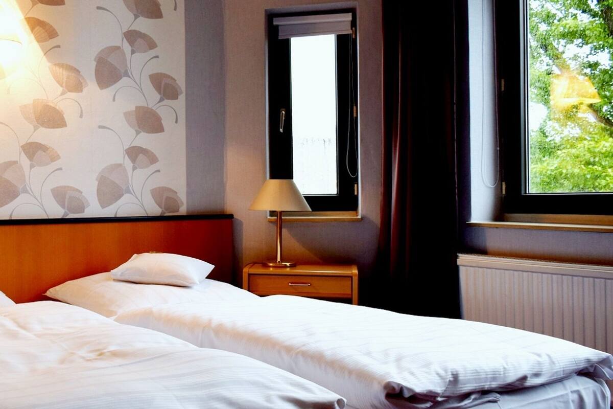 A hotel room with two single beds, a nightstand with a lamp, a wall-mounted light fixture, a window with a view of greenery, and a radiator.