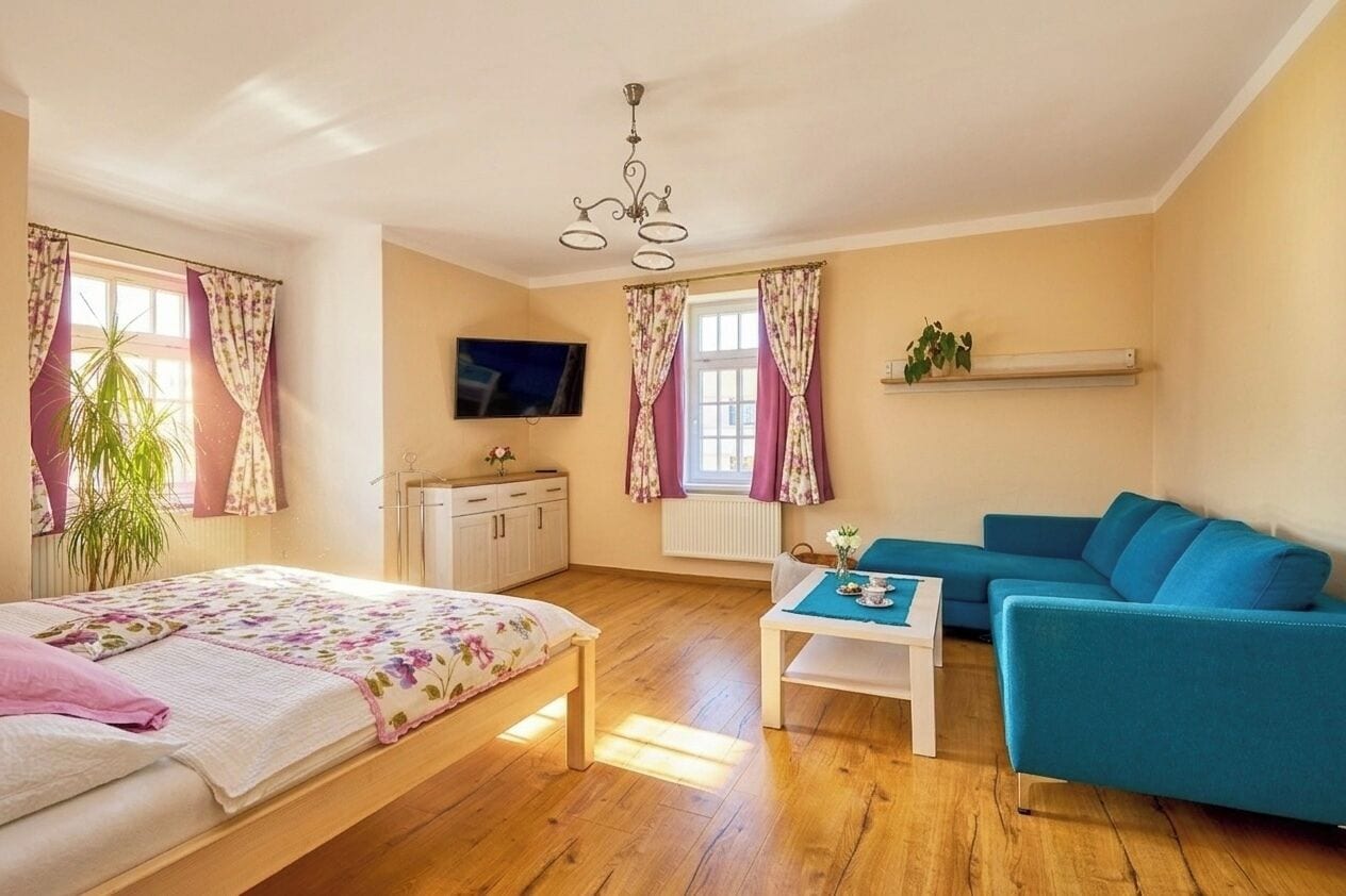 Un dormitorio con cama, sofá azul, mesita pequeña, televisor y ventana con cortinas.