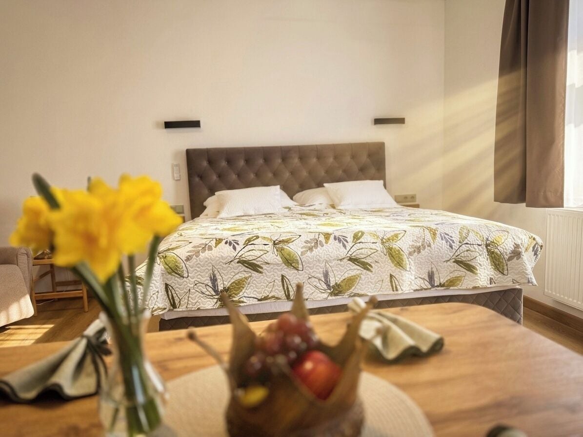 Un dormitorio con una cama, una mesita de noche con un florero y una ventana con cortinas.