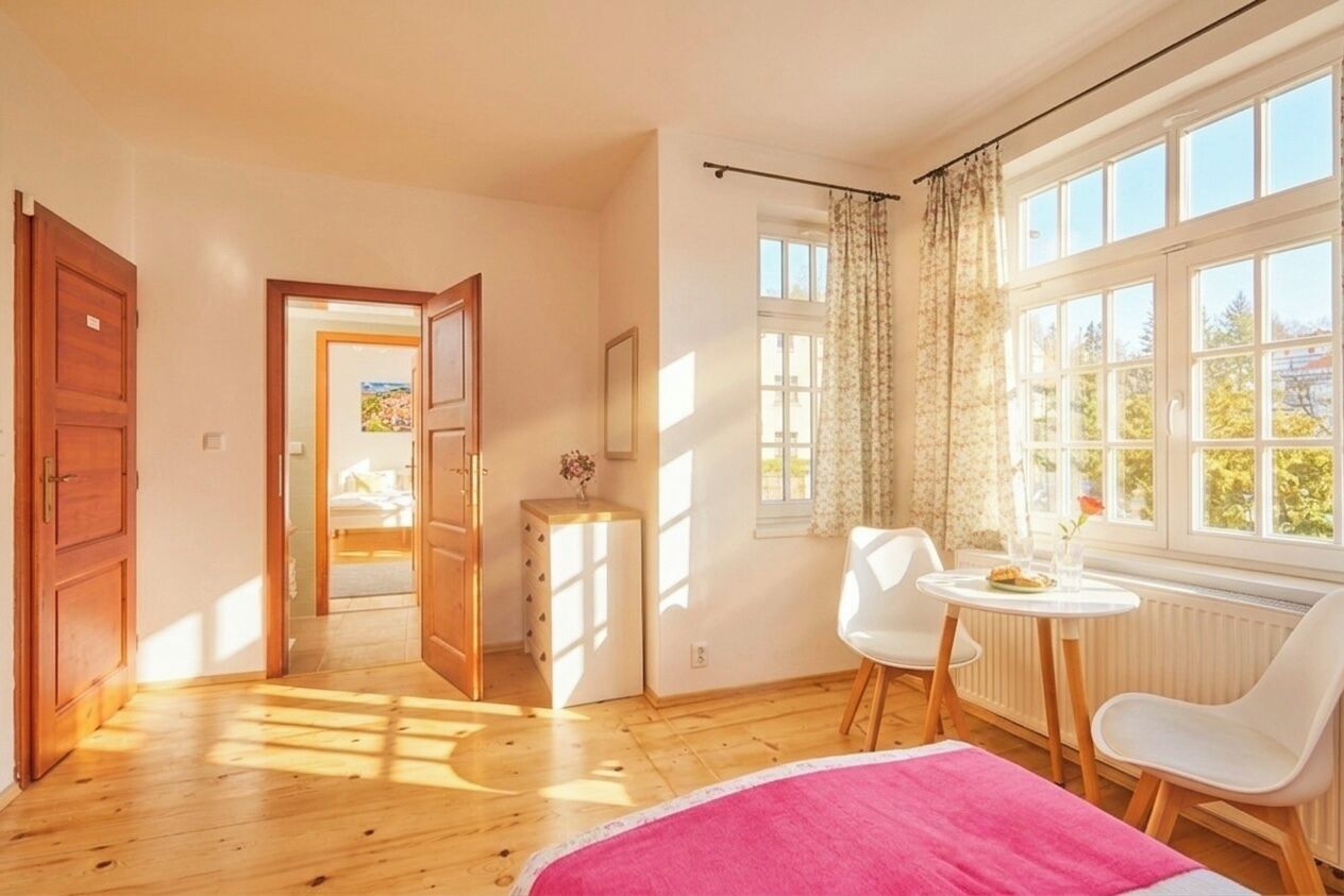 Une pièce comprenant un lit, une petite table avec des chaises, une fenêtre avec des rideaux et une porte donnant sur une autre pièce.