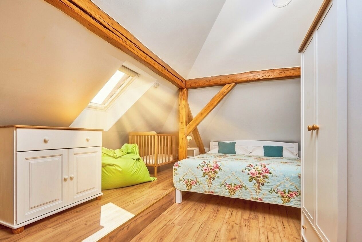 Un dormitorio con techo inclinado, vigas de madera, una cama con ropa de cama floral, un sillón de cuero en forma de saco y un aparador blanco.