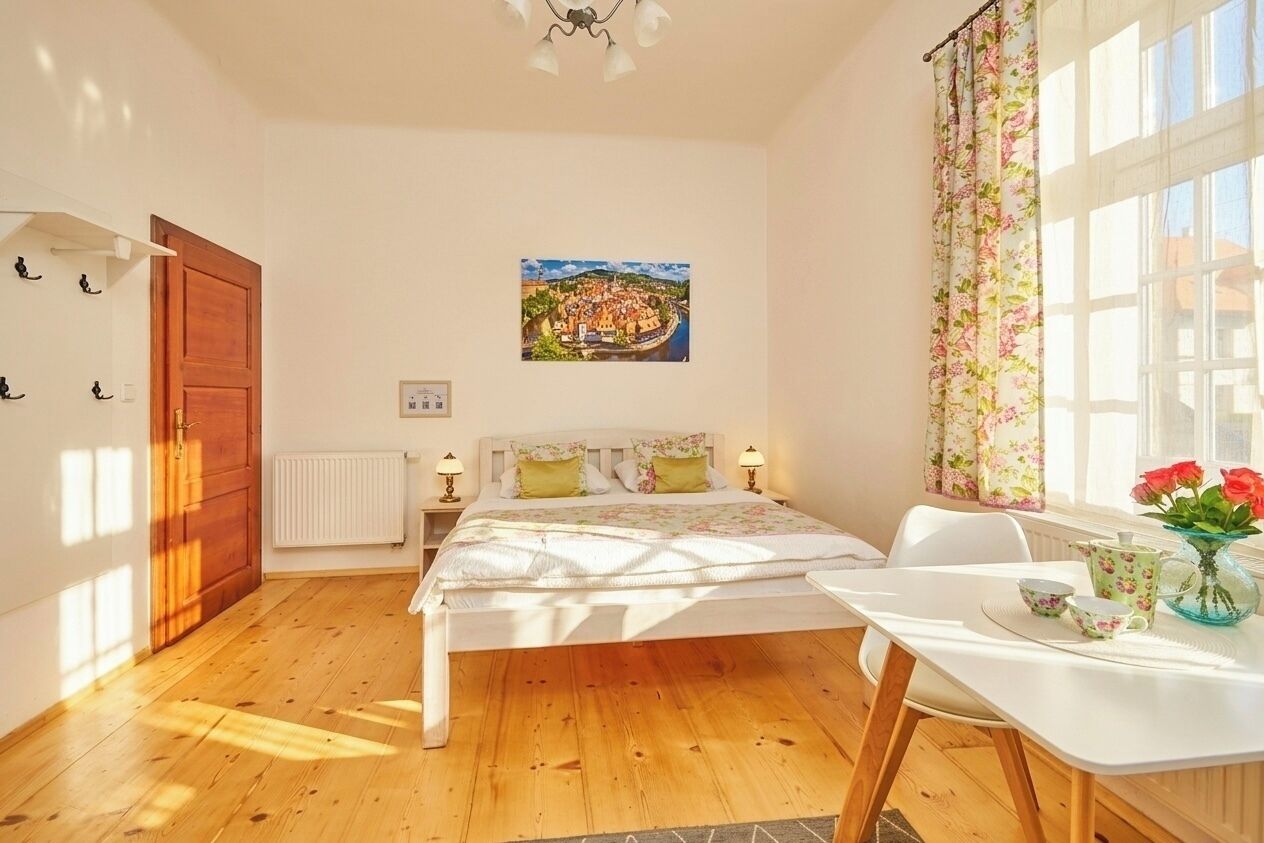 A bedroom with a wooden floor, a bed with floral bedding, a table with a vase of flowers, and a window with curtains.