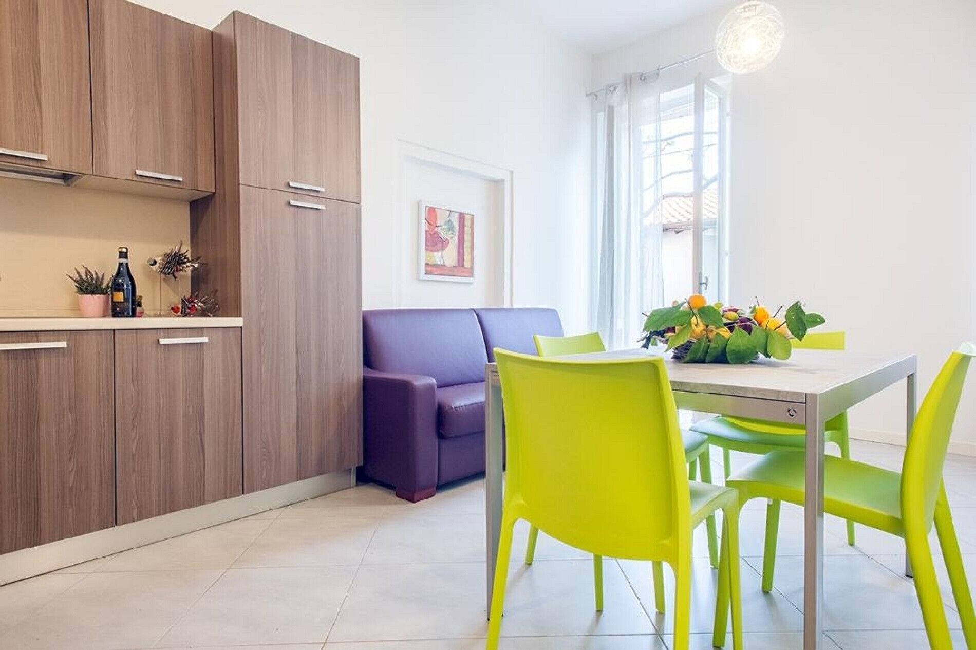 A modern kitchen with wooden cabinets, a purple sofa, and a dining area with a table and bright green chairs.