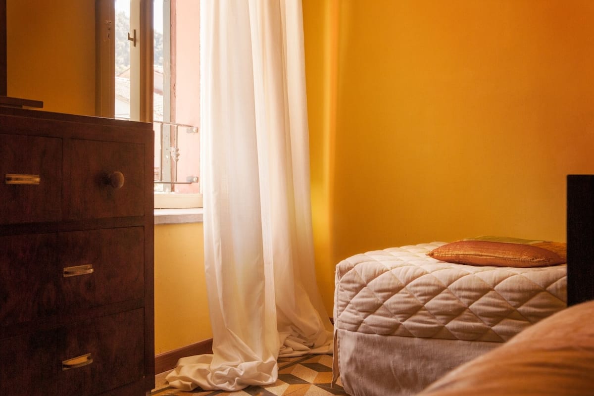 A room with a wooden dresser, a bed with a quilted cover, and a window with white curtains.