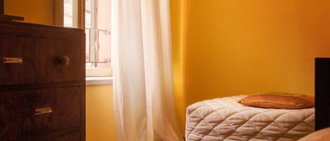 Una habitación con un aparador de madera, una cama con cubrecama acolchado y una ventana con cortinas blancas.