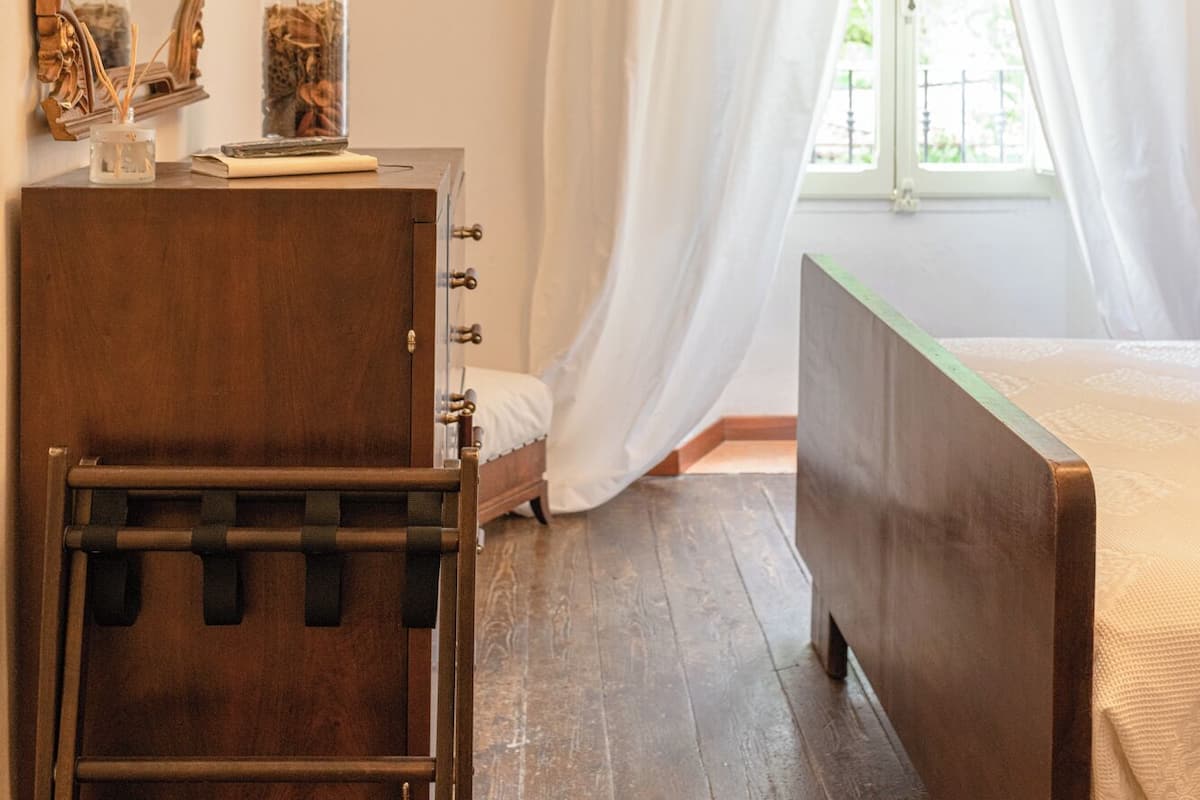 A bedroom with a wooden dresser, a bed, a TV mounted on the wall, and a window with white curtains.