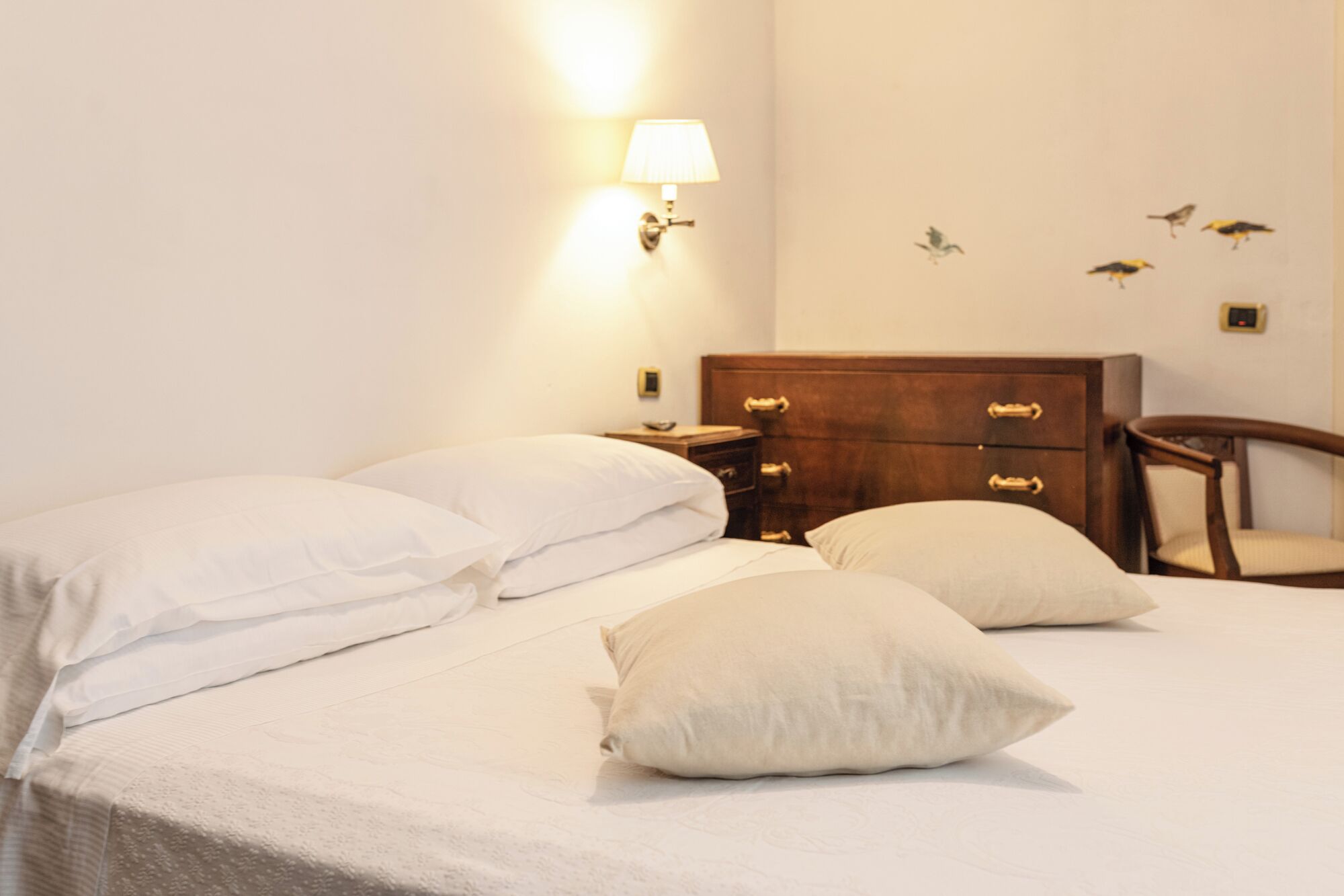 Una cama bien hecha con sábanas y almohadas blancas, una mesita de noche de madera con lámpara, y un aparador de madera con cajones.