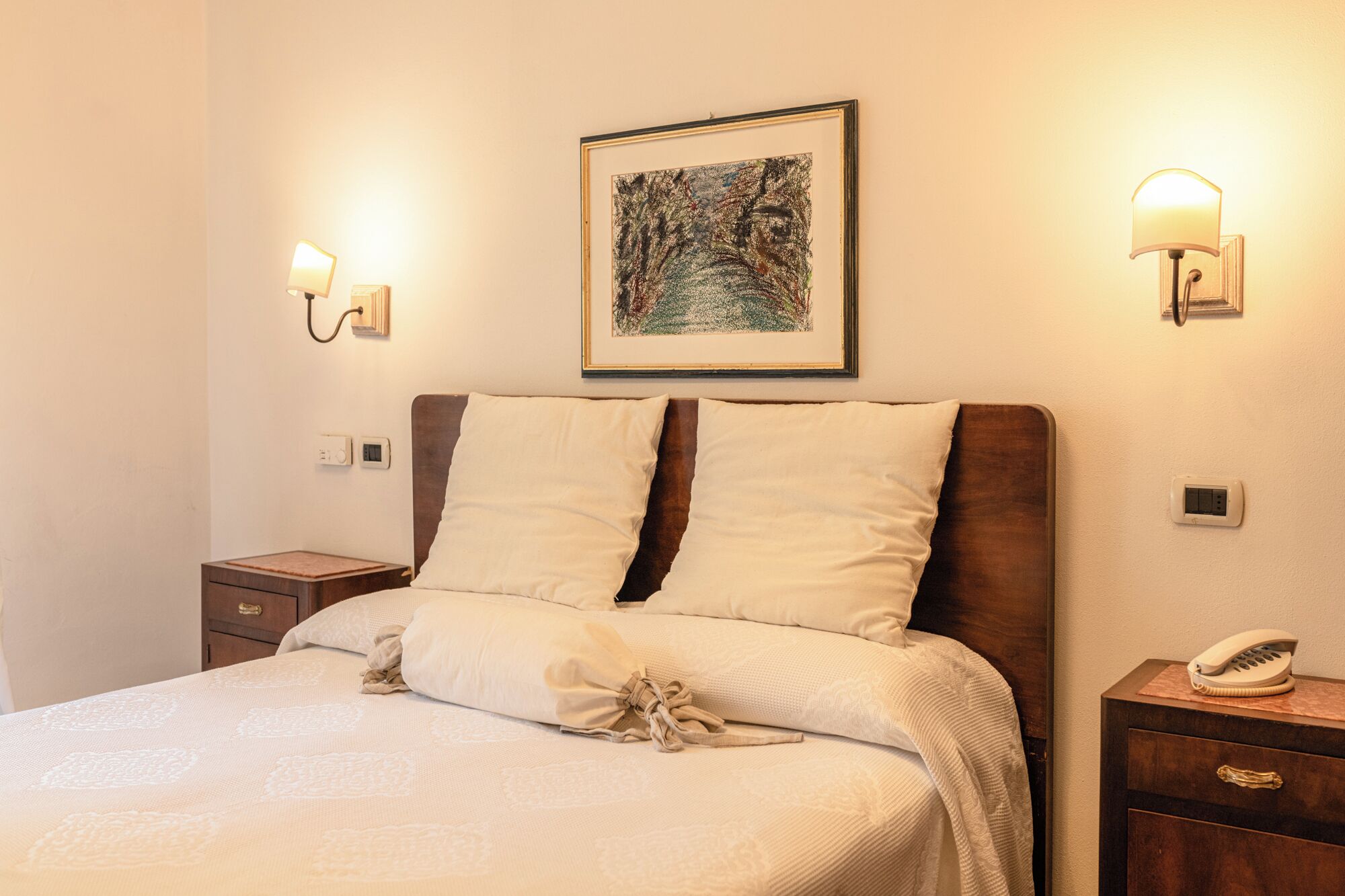 Una cama bien hecha con cabecera, dos mesitas de noche, un teléfono de pared y dos apliques.
