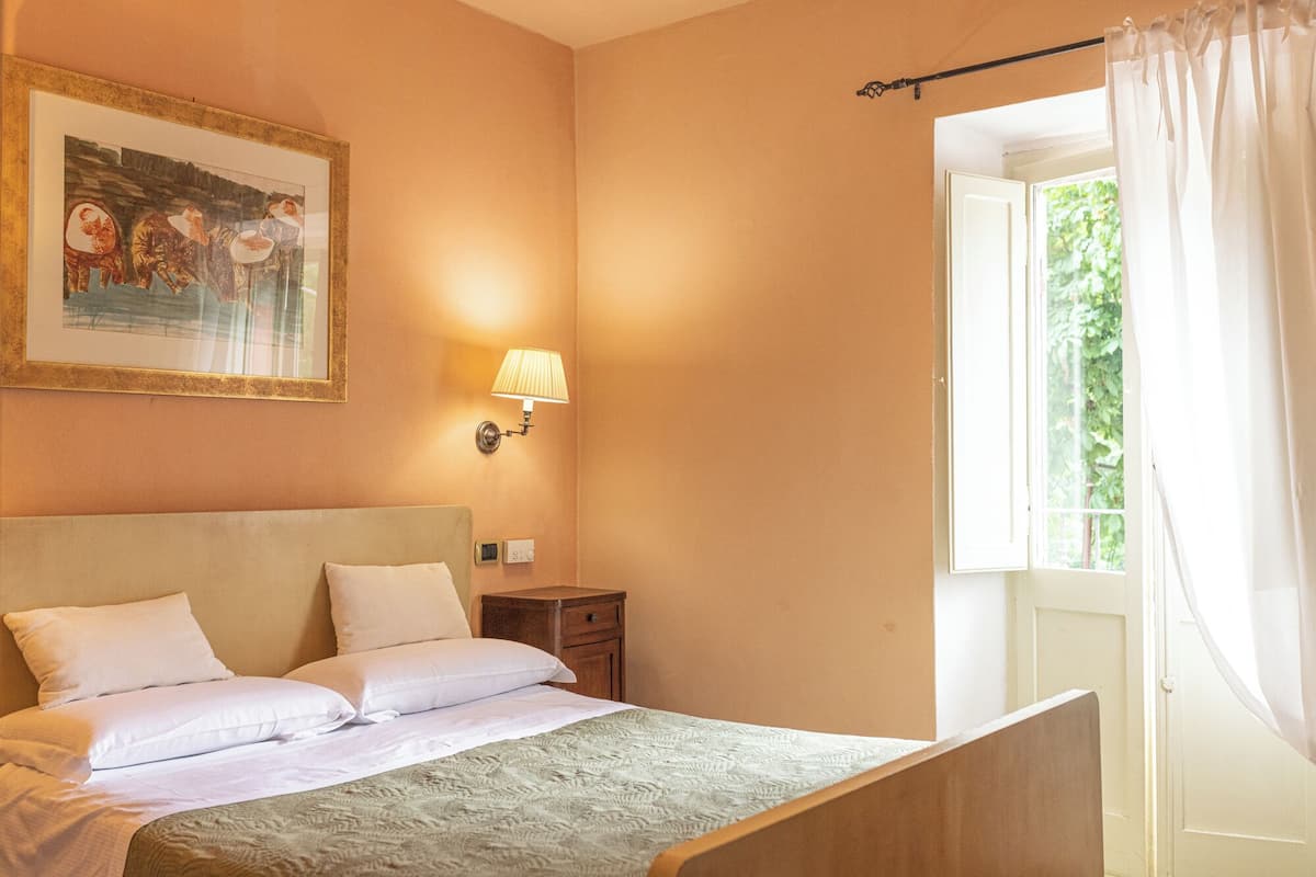 A bedroom with a bed, a nightstand, a lamp, a painting, and a window with curtains.