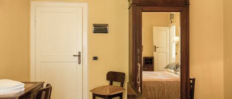 A hotel room with a bed, a desk with a chair, a wardrobe, a mirror, and a door.
