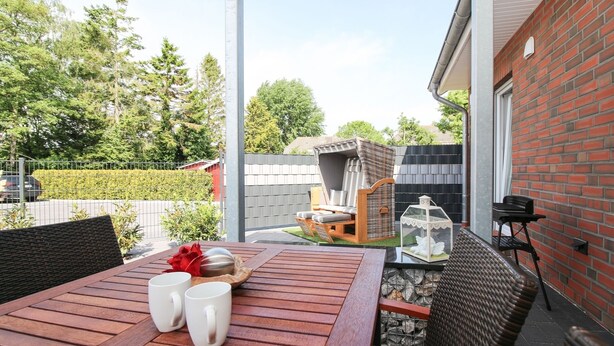 Eine Terrasse mit einem Holztisch, zwei weißen Tassen und einem Korbstuhl.