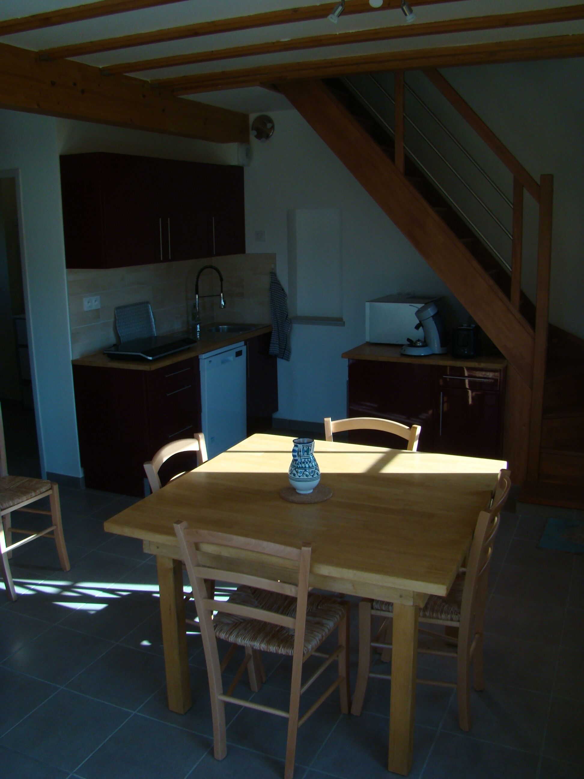 Una cocina con armarios de madera, un lavamanos y una cafetera. Hay una mesa de comedor con sillas y un florero encima.