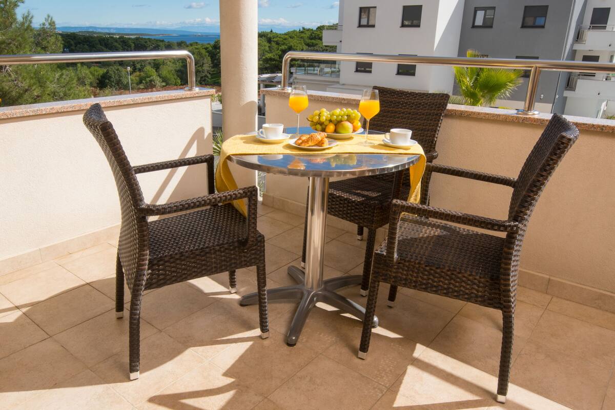 A balcony with wicker chairs, a table with breakfast items, and a view of the sea and buildings.