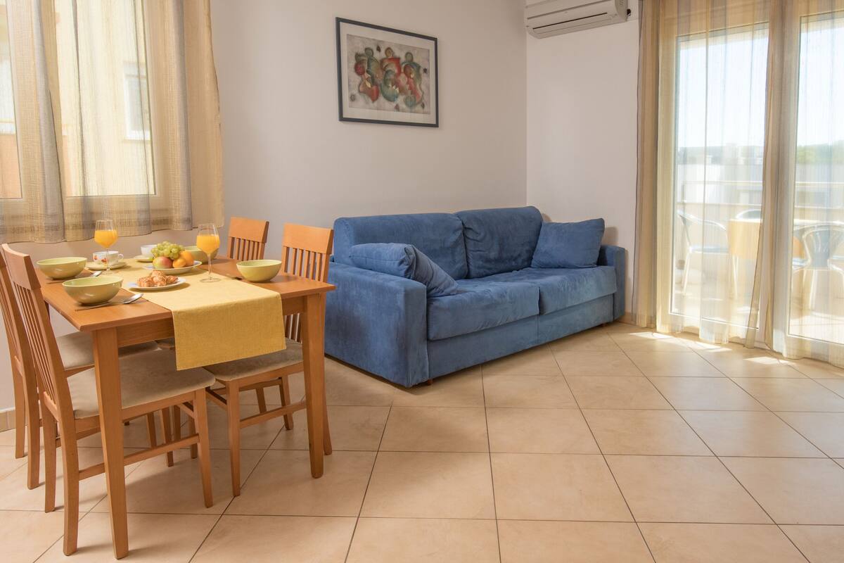 A dining area with a table set for two, a blue sofa, and a window with curtains.