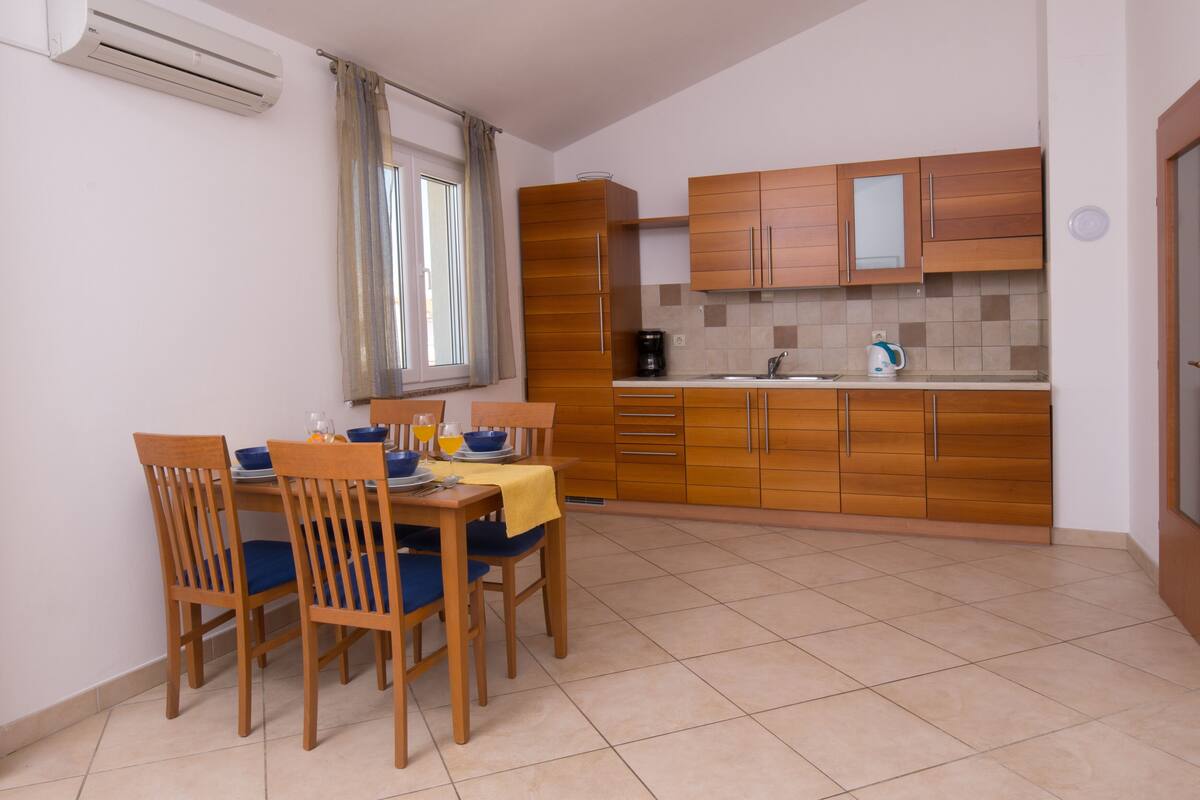 A compact kitchen with wooden cabinets, a dining table with chairs, and a window with curtains.