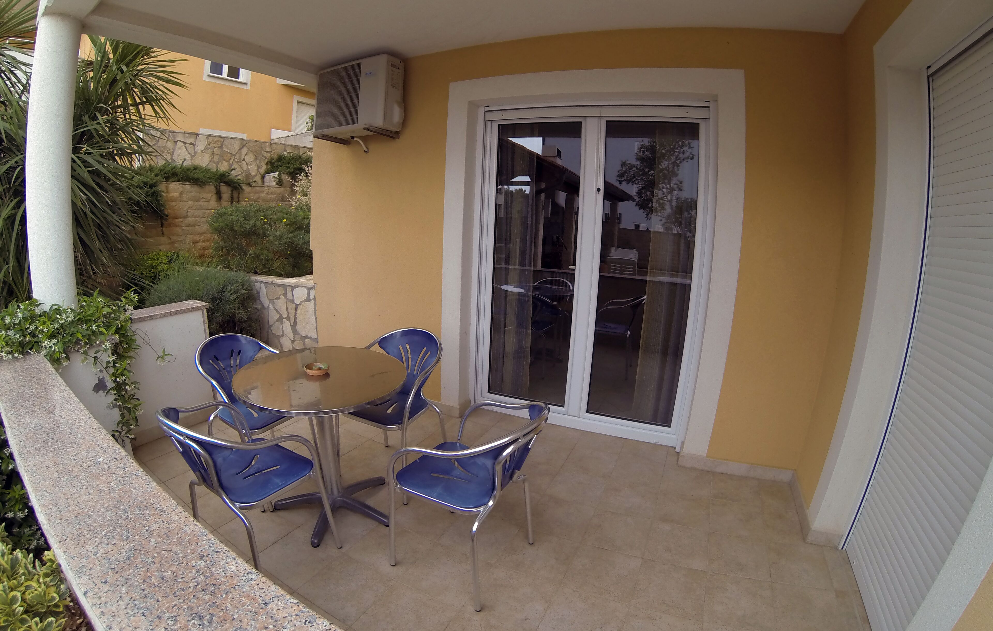 Eine Terrasse mit rundem Tisch und vier blauen Stühlen, einer Schiebetür und einer Steinwand mit Grünpflanzen.
