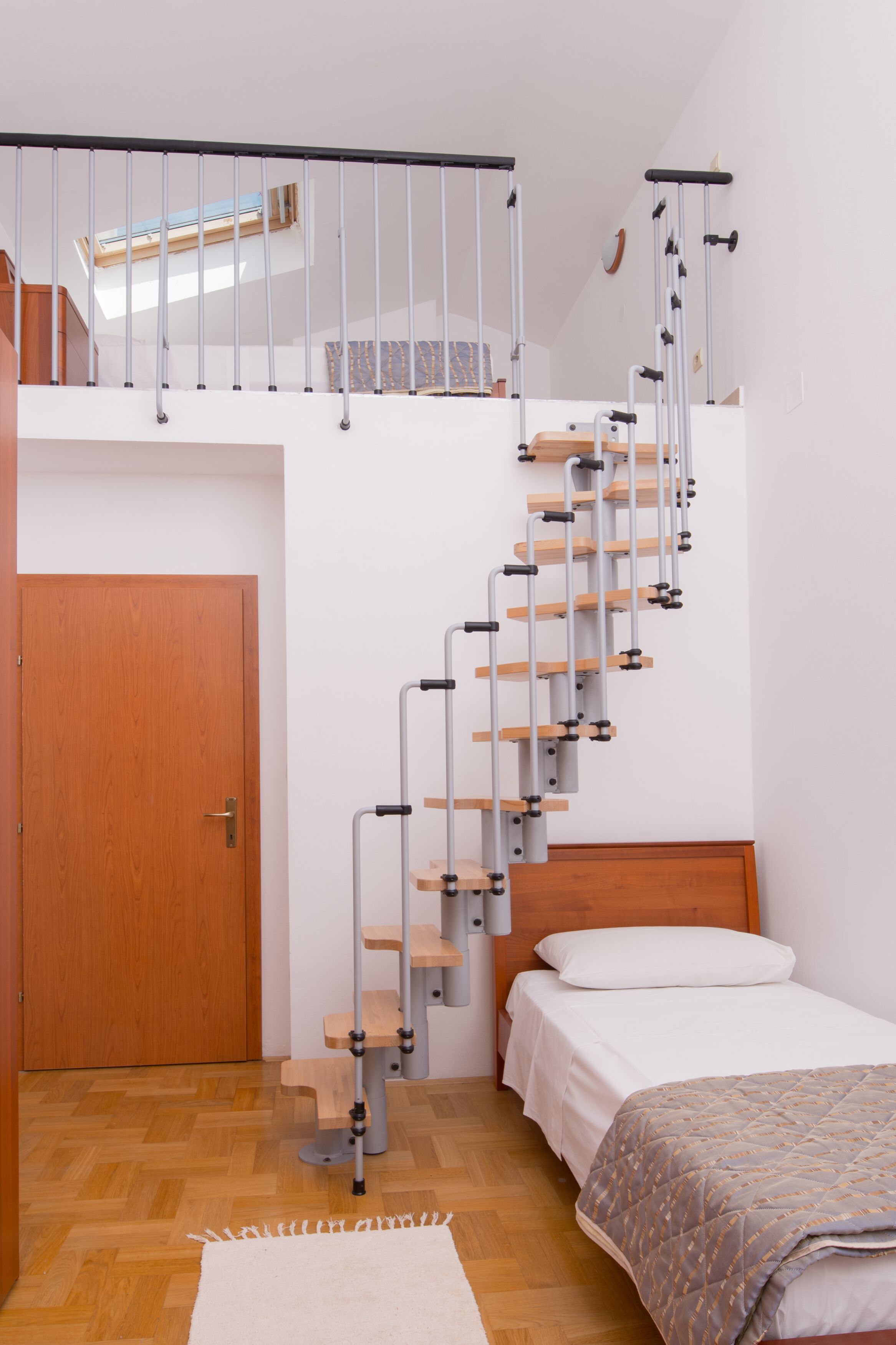 Une chambre avec un lit en bois, un mur blanc, une porte en bois et un escalier avec des rampes en métal.