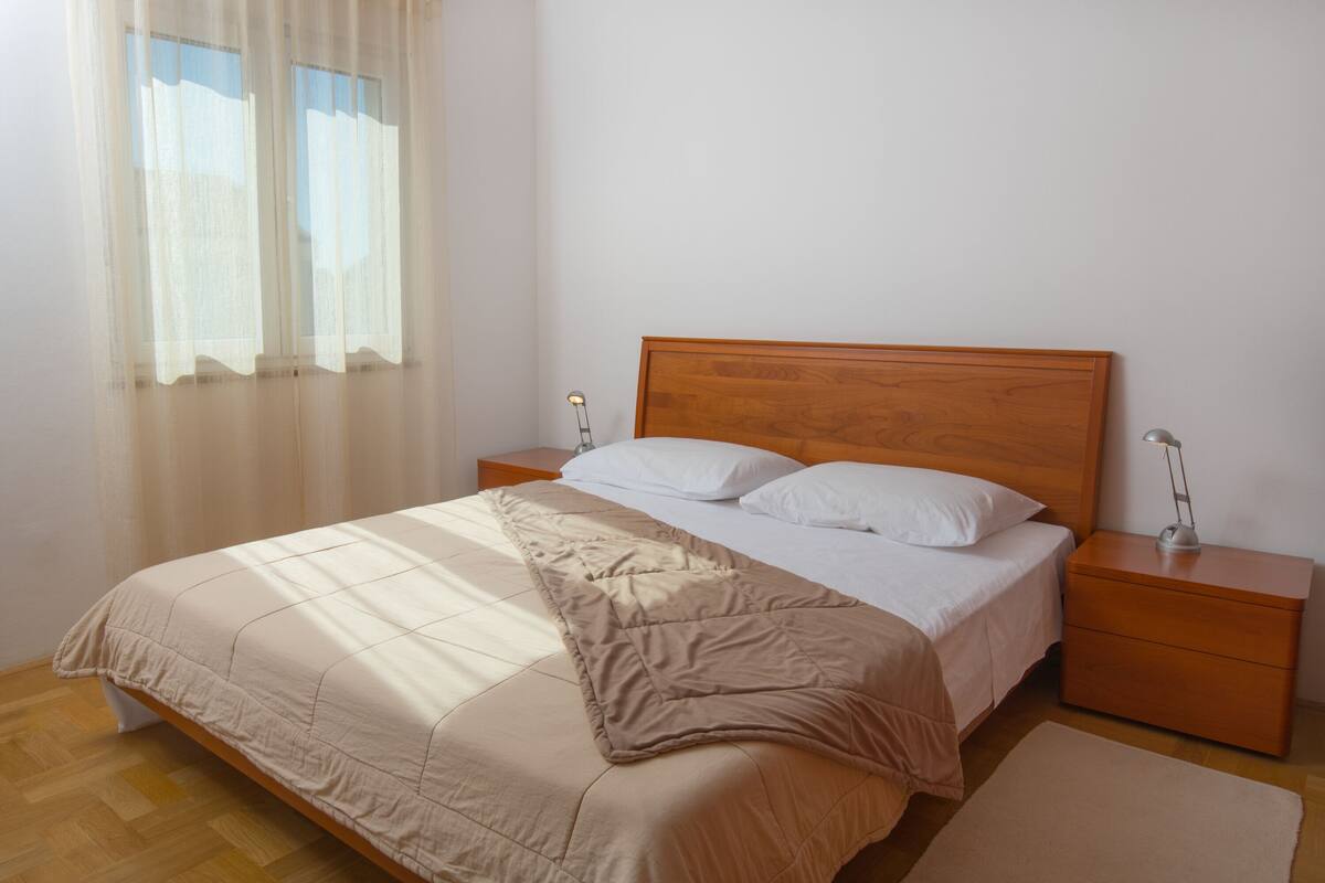 A bedroom with a wooden bed, white bedding, two bedside tables with lamps, and a window with sheer curtains.