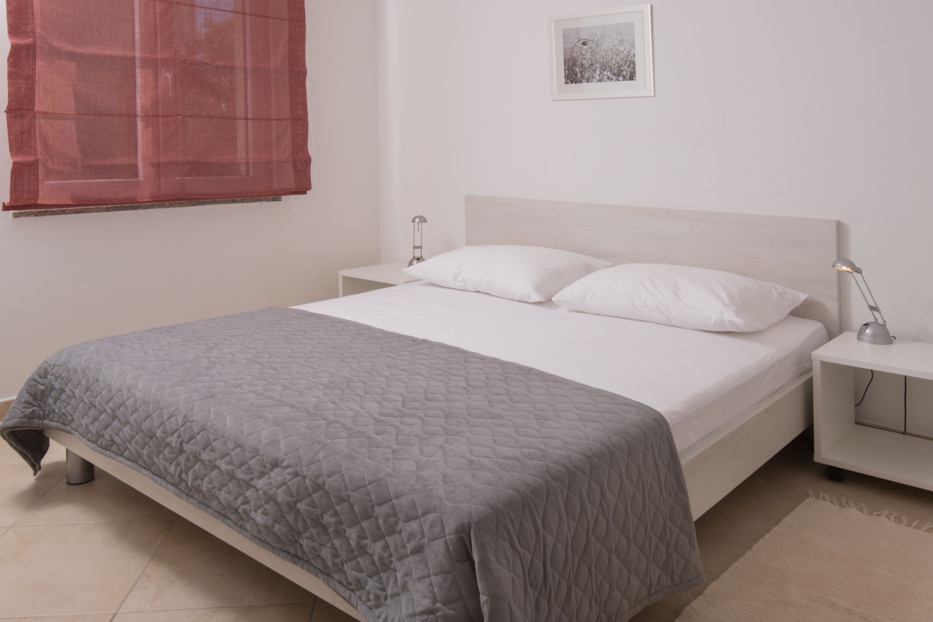 Un letto rifatto con cura, con una coperta grigia, cuscini bianchi e una testata bianca. È presente un comodino con una lampada sopra.