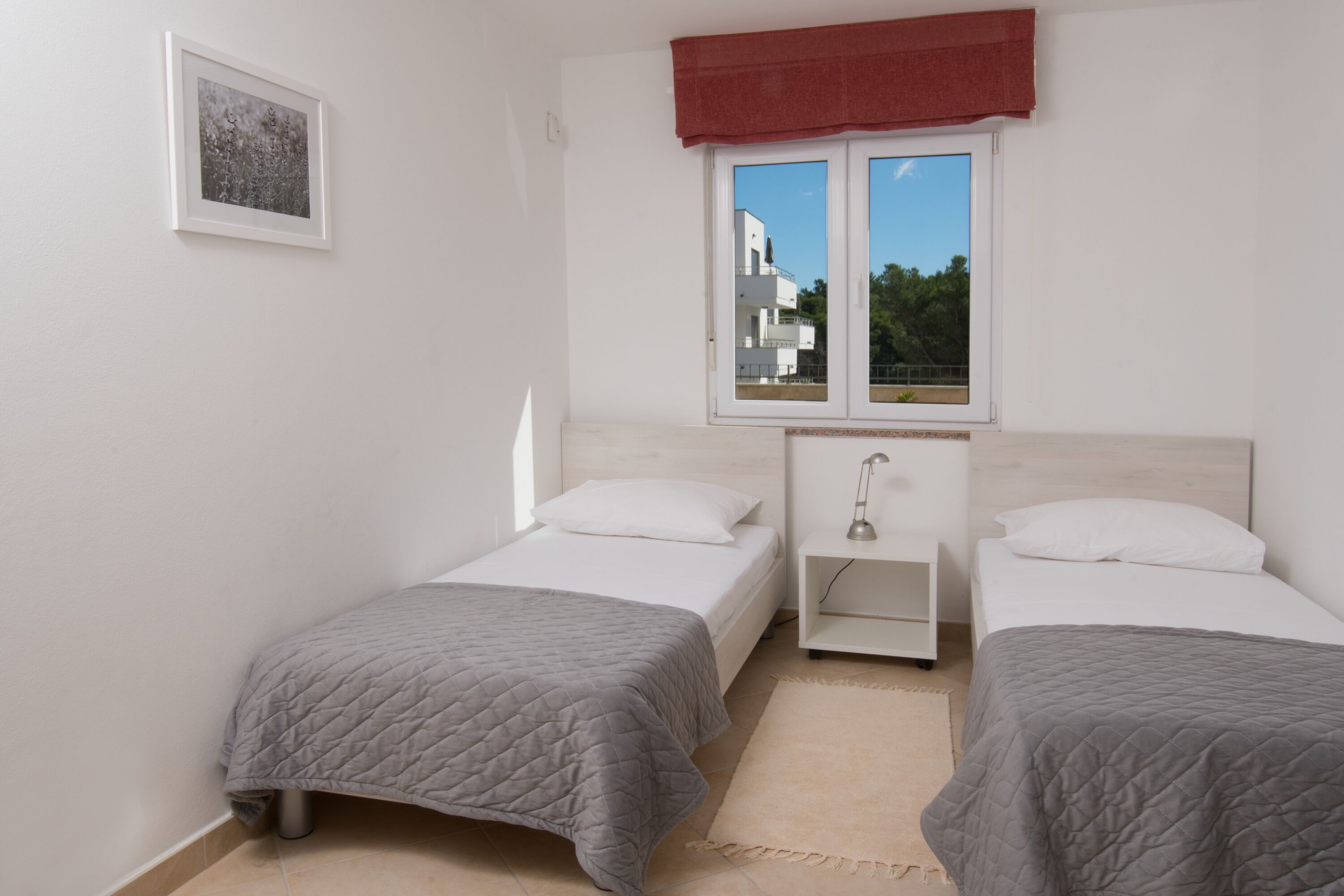 A bedroom with two beds, a window with a view of buildings, a small bedside table, and a framed picture on the wall.