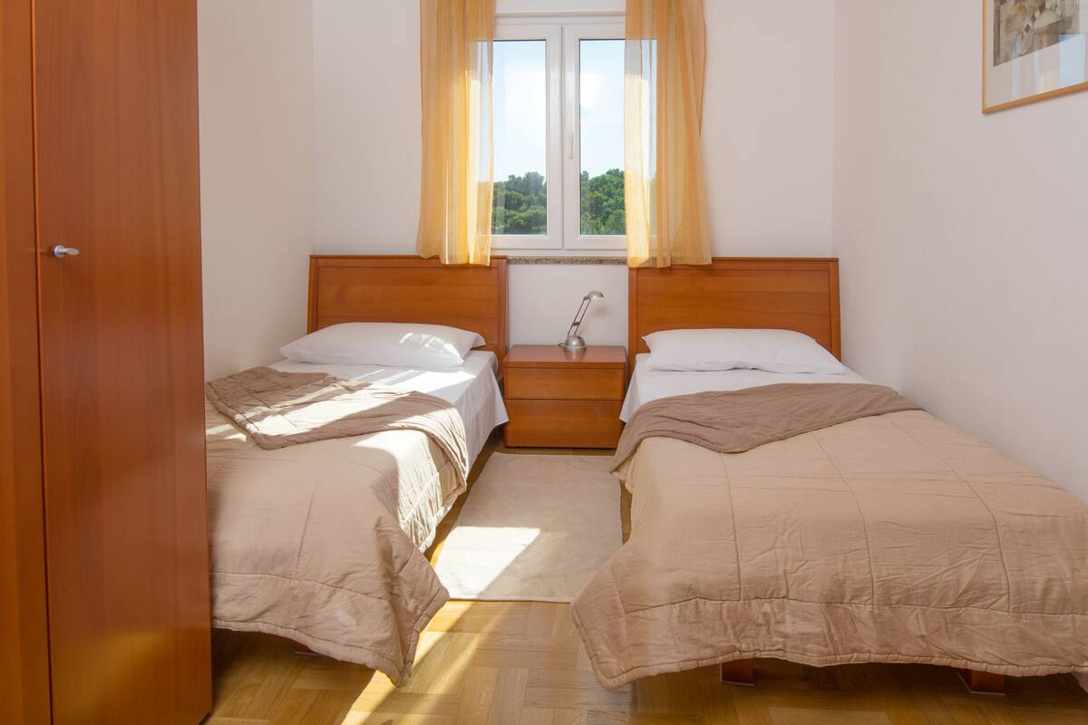 A bedroom with two beds, a wooden headboard, a nightstand with a lamp, and a window with curtains.