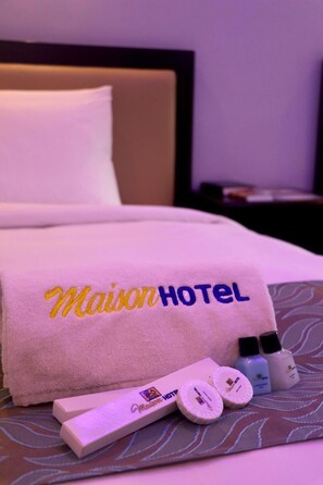 Uma cama de hotel com uma toalha Maison Hotel, uma caixa de produtos de higiene e duas garrafas de loção.